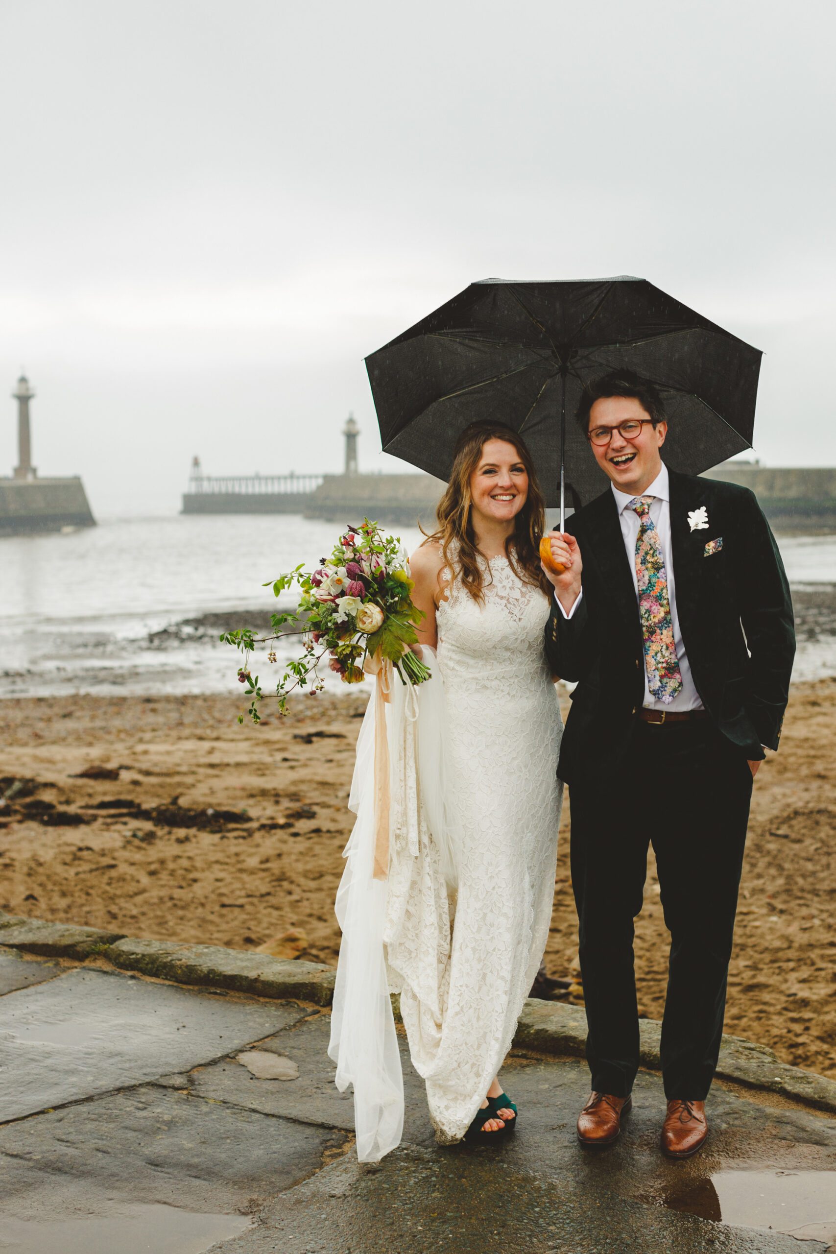 Bride and groom standing with an umbrella at Whitby beach on their rainy day wedding. Flowers by Moonwind Flowers.